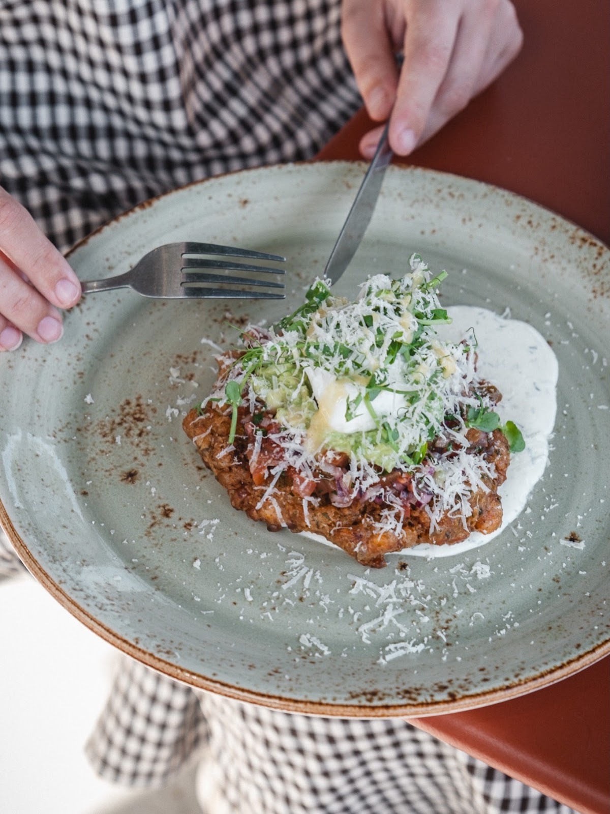 A fritter topped with grated hard cheese, avocado, chopped tomato and pea shoots, on a green speckled plate held over the table, knife and fork in hand.