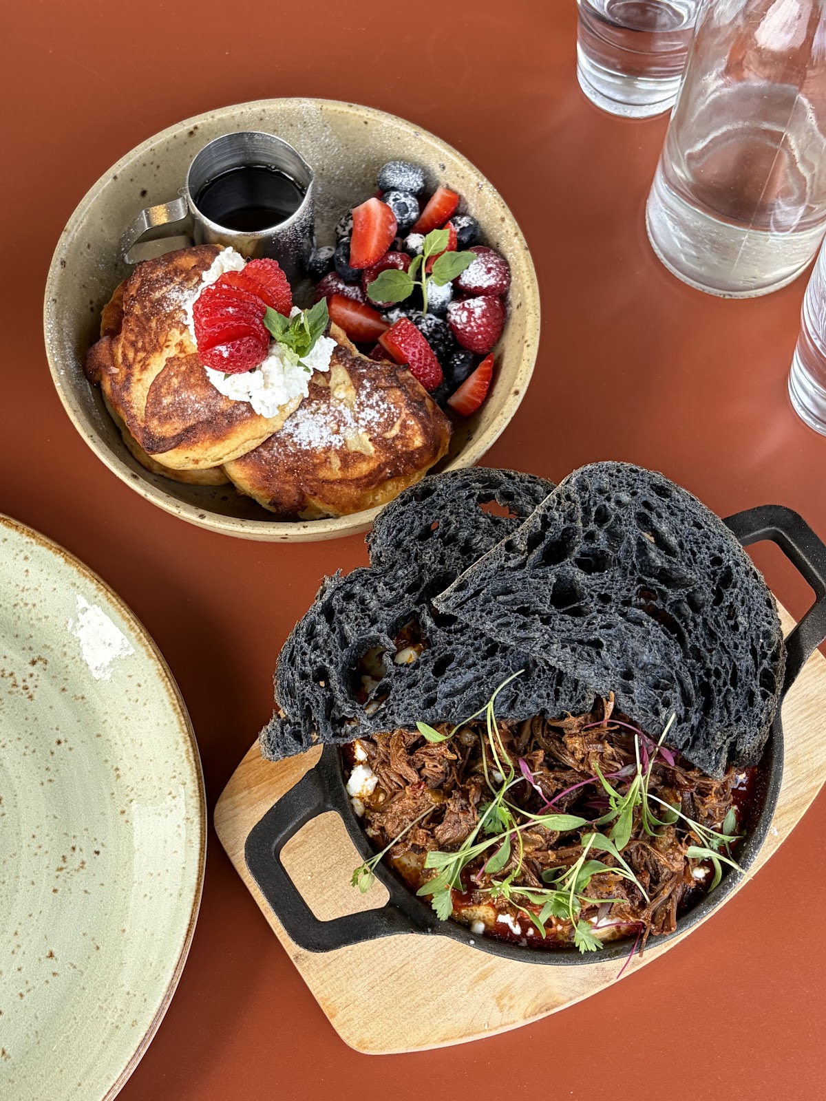 Seen from above on a terracotta table: a bowl of thick pancakes with berries, cream and a jug of syrup, beside a cast-iron pan of slow-cooked shredded meat topped with herbs and two slices of charcoal-black sourdough.