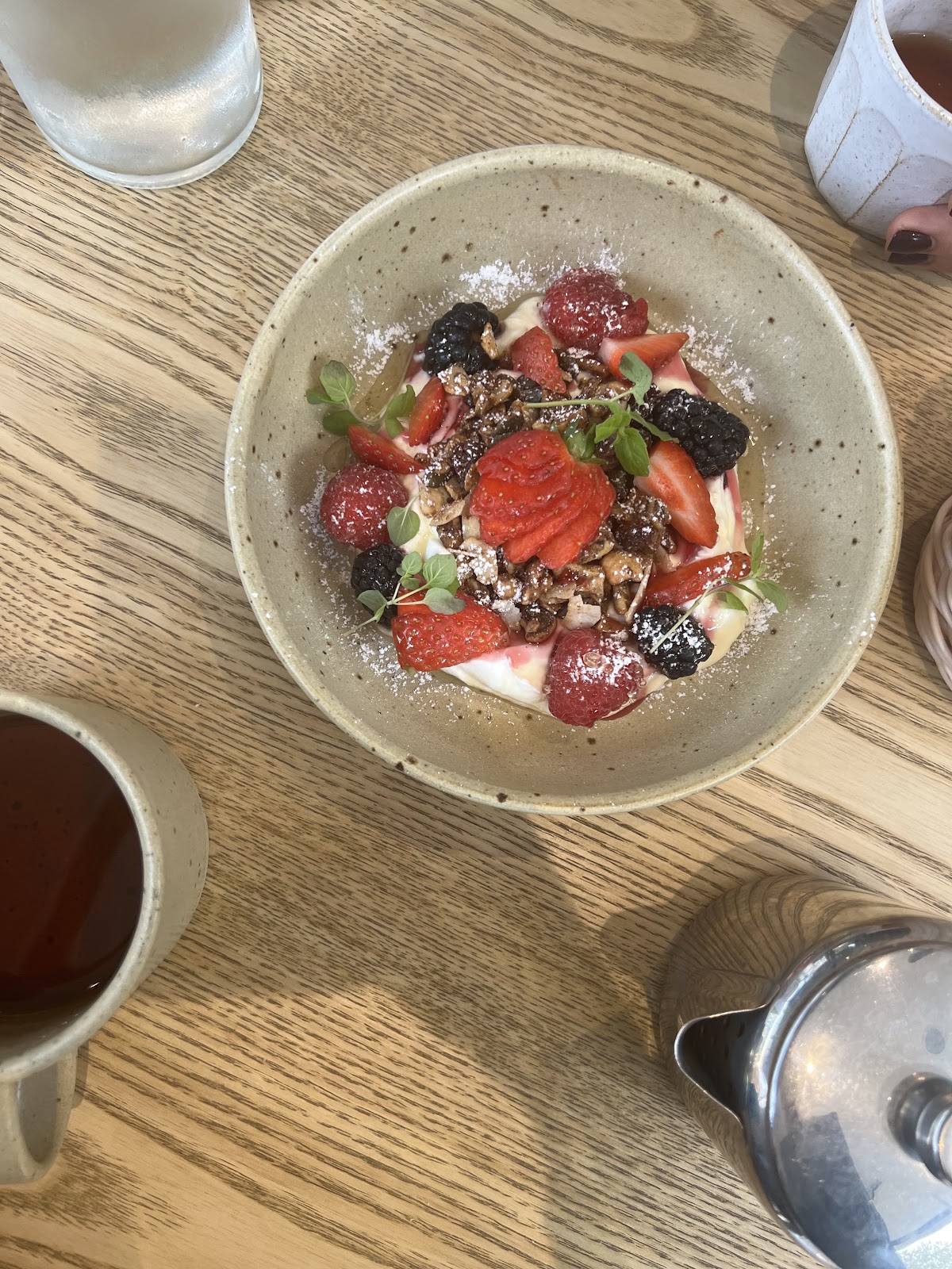 A speckled stoneware bowl of yoghurt, granola, strawberries, raspberries and blackberries dusted with icing sugar, on a pale oak table with a cup of tea alongside.