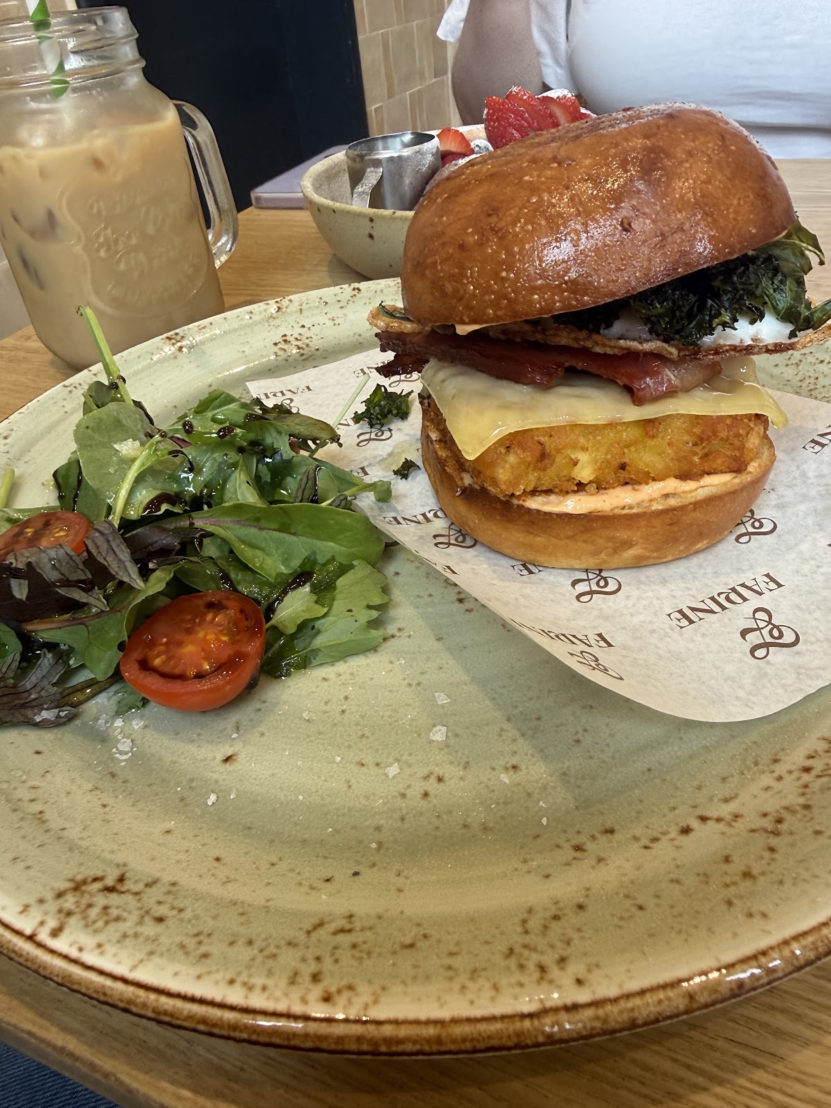 A glazed bun filled with a potato rösti, melted cheese, bacon, egg and crisp greens, served on paper printed FARINE beside a dressed salad, with an iced coffee in a glass jar behind.