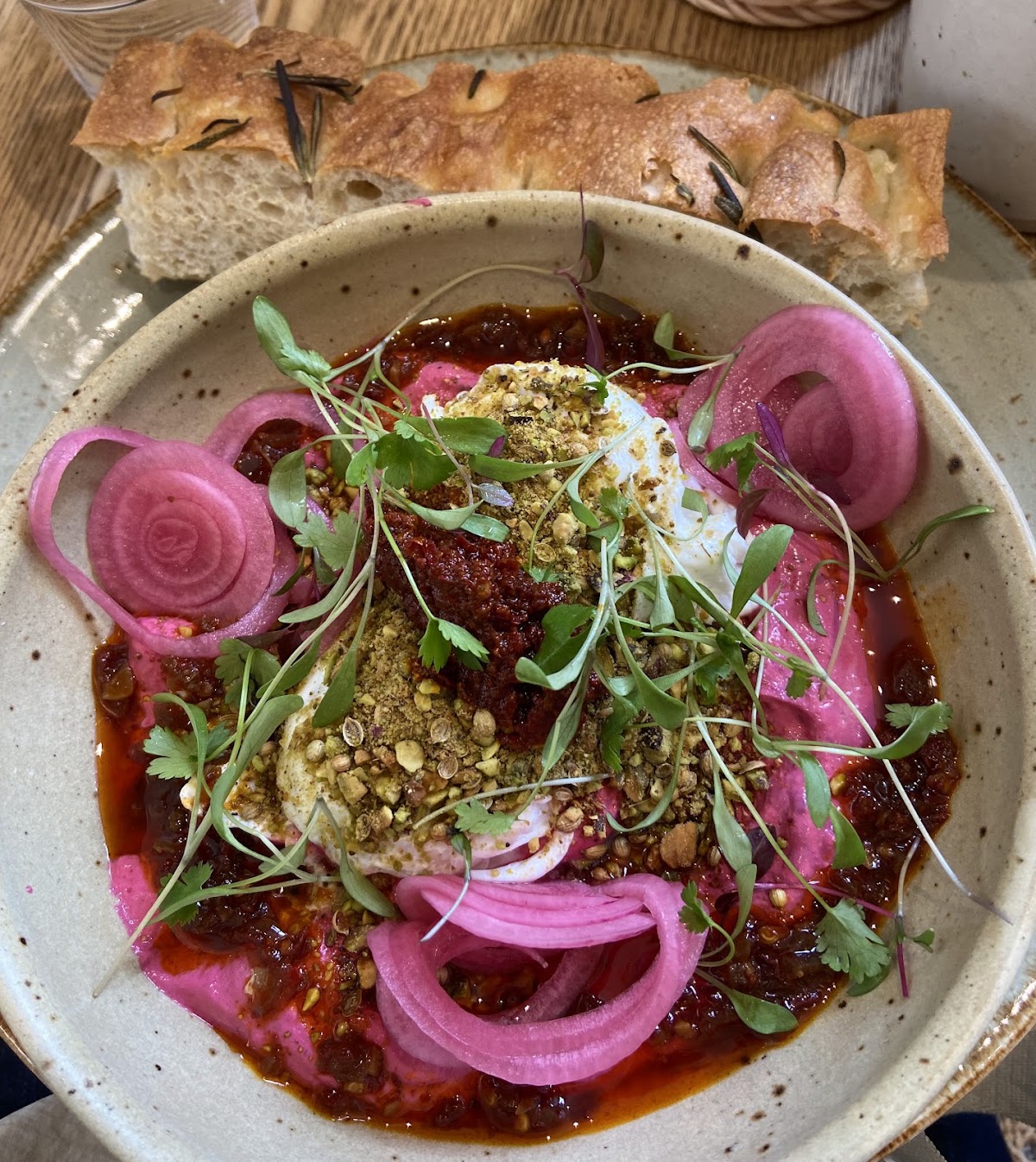 A wide bowl of whipped white cheese over bright pink beetroot purée with chilli oil, crushed pistachio, coriander and pickled onions, with rosemary focaccia behind.